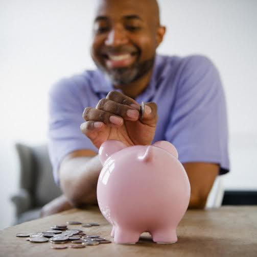 Smiling African American man putting coins in piggy bank --- Image by © JGI/Jamie Grill/Blend Images/Corbis